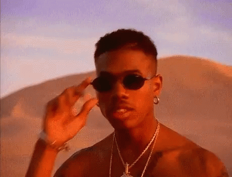 A Man Wearing Sunglasses And A Necklace Is Standing In The Desert . GIF