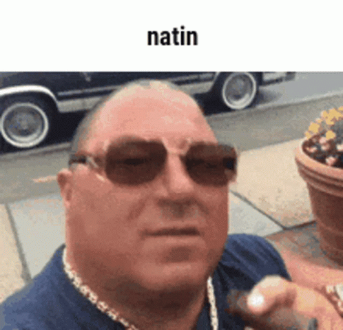 A Man Wearing Sunglasses And A Necklace Is Standing On A Sidewalk In Front Of A Car . GIF