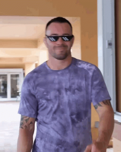 A Man Wearing Sunglasses And A Purple Tie Dye Shirt Is Smiling GIF