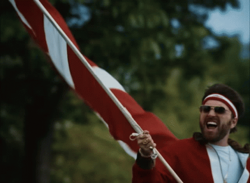 A Man Wearing Sunglasses And A Red Jacket Is Holding A Red And White Flag GIF