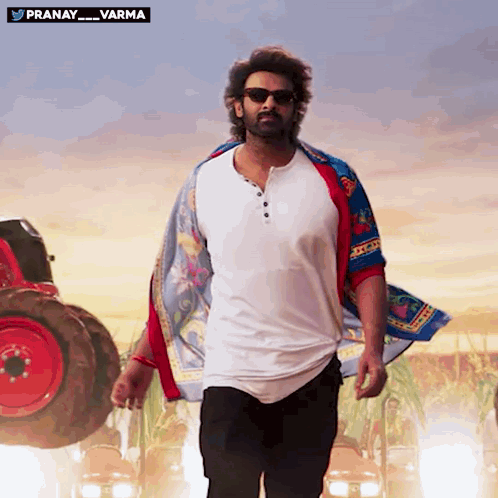 A Man Wearing Sunglasses And A Scarf Is Walking In Front Of A Red Tractor With The Words Pranay Varma Above Him GIF