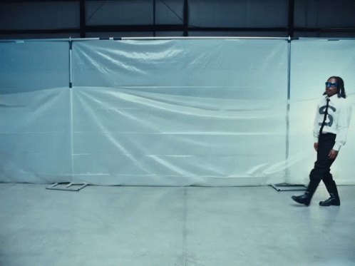 A Man Wearing Sunglasses And A Shirt With A Dollar Sign On It Stands In Front Of A White Tarp GIF