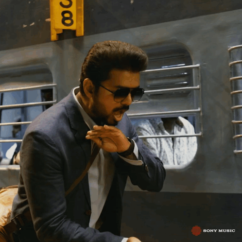 A Man Wearing Sunglasses And A Suit Is Standing In Front Of A Yellow Sign That Says Sony Music GIF
