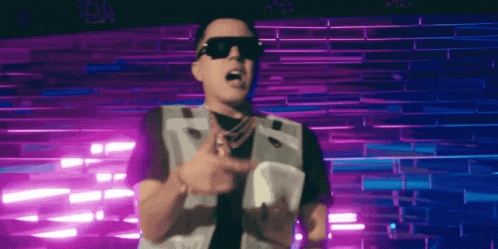 A Man Wearing Sunglasses And A Vest Stands In Front Of A Purple Wall GIF