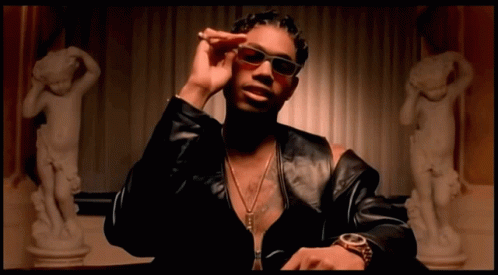 A Man Wearing Sunglasses And A Watch Is Sitting In Front Of A Statue Of A Woman GIF