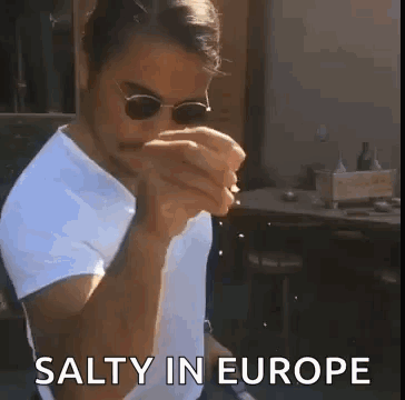 A Man Wearing Sunglasses And A White Shirt Is Eating A Sandwich While Sitting At A Table . GIF