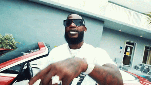 A Man Wearing Sunglasses And A White Shirt Is Standing In Front Of A Red Car GIF