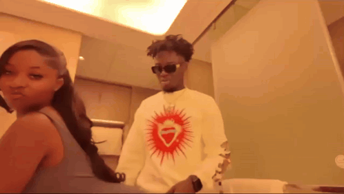A Man Wearing Sunglasses And A White Shirt With A Red Heart On It Stands Next To A Woman GIF