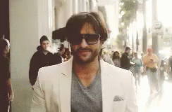 A Man Wearing Sunglasses And A White Suit Is Walking Down A Street . GIF
