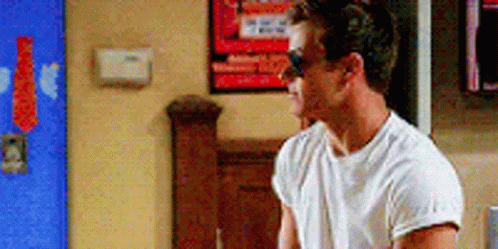 A Man Wearing Sunglasses And A White T-Shirt Is Standing In A Room . GIF