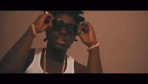 A Man Wearing Sunglasses And A White Tank Top Has Gold Chains Around His Neck GIF