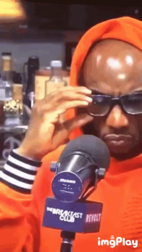 A Man Wearing Sunglasses And An Orange Hoodie Stands In Front Of A Breakfast Club Microphone GIF