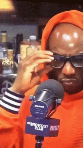 A Man Wearing Sunglasses And An Orange Hoodie Stands In Front Of A Microphone That Says The Breakfast Club GIF