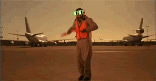 A Man Wearing Sunglasses And An Orange Vest Stands In Front Of Planes GIF