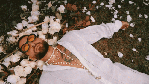 A Man Wearing Sunglasses And Pearls Is Laying In The Grass Surrounded By Flowers GIF