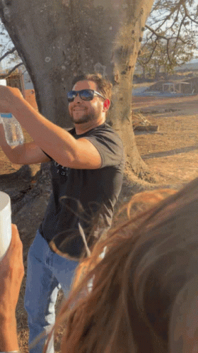 A Man Wearing Sunglasses Holds A Bottle Of Water In Front Of A Tree GIF