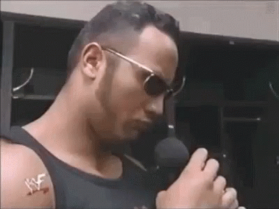 A Man Wearing Sunglasses Is Holding A Microphone And Talking Into It . GIF