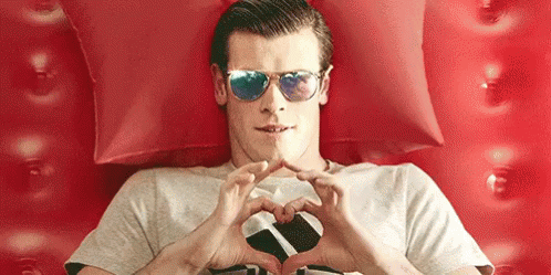 A Man Wearing Sunglasses Is Making A Heart Shape With His Hands While Laying On A Bed . GIF