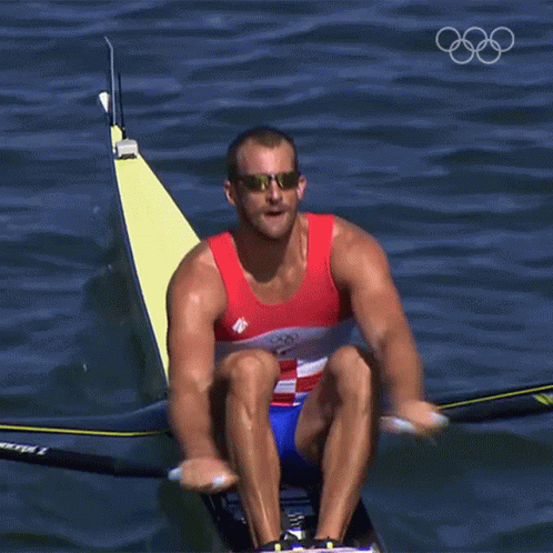 A Man Wearing Sunglasses Is Rowing A Boat In The Water With The Olympic Rings In The Background GIF