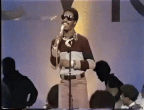 A Man Wearing Sunglasses Is Singing Into A Microphone While Standing In Front Of A Crowd . GIF