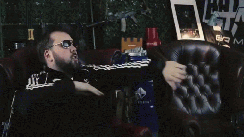 A Man Wearing Sunglasses Is Sitting In A Chair In Front Of A Sign That Says Am GIF