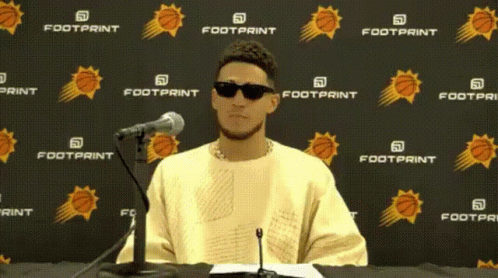 A Man Wearing Sunglasses Is Sitting In Front Of A Footprint Banner GIF