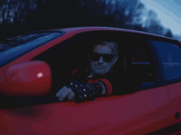 A Man Wearing Sunglasses Is Sitting In The Driver 'S Seat Of A Red Car GIF