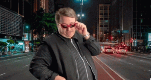 A Man Wearing Sunglasses Is Standing In Front Of A City Street At Night GIF