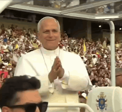 A Man Wearing Sunglasses Is Standing In Front Of A Crowd Of People Applauding The Pope . GIF
