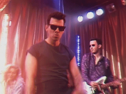 A Man Wearing Sunglasses Is Standing On A Stage With A Guitar Player Behind Him . GIF