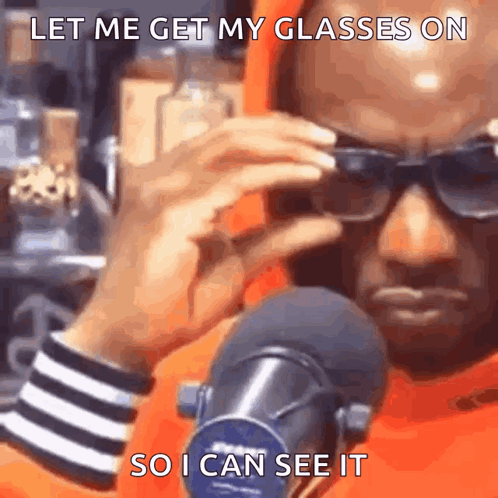 A Man Wearing Sunglasses Is Talking Into A Microphone While Holding His Glasses On . GIF
