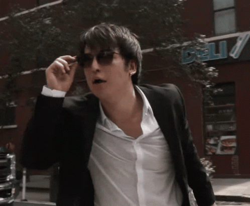A Man Wearing Sunglasses Is Walking Down The Street In Front Of A Deli GIF