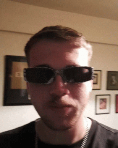 A Man Wearing Sunglasses Looks At The Camera In Front Of A Wall With Pictures On It GIF