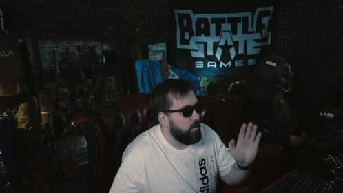 A Man Wearing Sunglasses Sits In Front Of A Battle State Games Sign GIF
