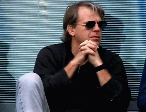 A Man Wearing Sunglasses Sits With His Hands Folded In Front Of A Window . GIF