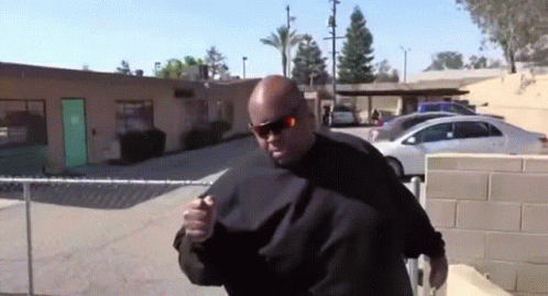 A Man Wearing Sunglasses Stands In Front Of A Building With A Green Door And Cars Parked In Front Of It GIF