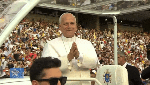 A Man Wearing Sunglasses Stands In Front Of A Crowd Of People While The Pope Applauds In The Background GIF