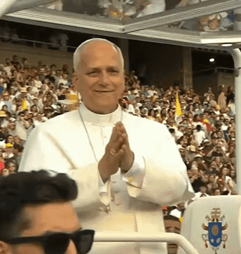 A Man Wearing Sunglasses Stands In Front Of A Crowd With A Shield That Says Papal GIF