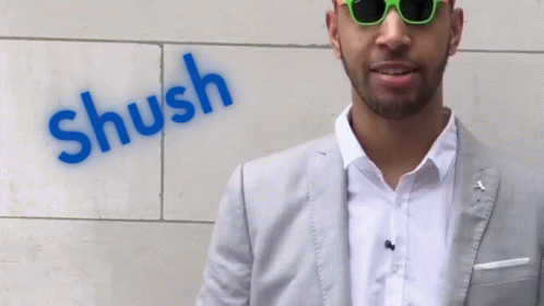 A Man Wearing Sunglasses Stands In Front Of A Wall That Has The Word Shush Painted On It GIF