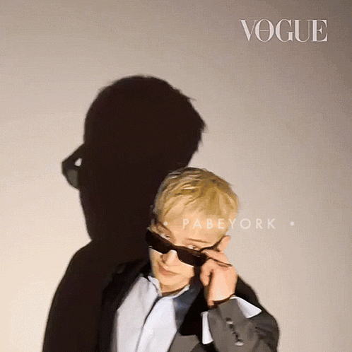 A Man Wearing Sunglasses Stands In Front Of A Wall That Says Vogue GIF