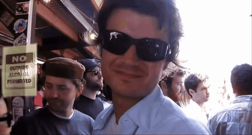 A Man Wearing Sunglasses Stands Next To A Sign That Says No Outside Alcohol Permitted GIF