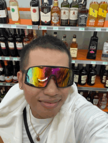 a man wearing sunglasses takes a selfie in front of a shelf of alcohol including jack daniel 's