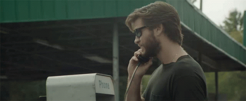 A Man Wearing Sunglasses Talks On A Pay Phone GIF