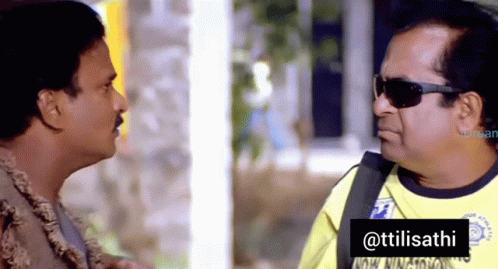 A Man Wearing Sunglasses Talks To Another Man In A Yellow Shirt That Says ' Ttilisathi ' On It GIF