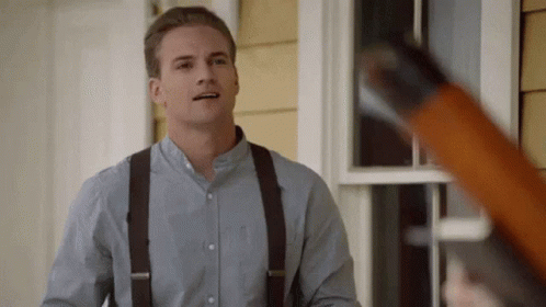 A Man Wearing Suspenders And A Blue Shirt Is Standing In Front Of A House Holding A Shotgun . GIF