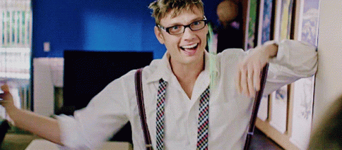 A Man Wearing Suspenders And Glasses Is Smiling And Leaning Against A Wall GIF