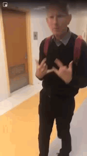 A Man With A Backpack Is Standing In A Hallway Making A Peace Sign With His Hands . GIF