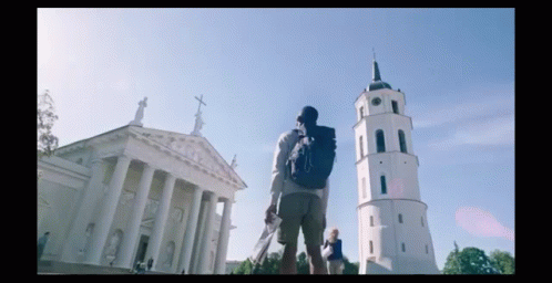 A Man With A Backpack Stands In Front Of A Large Building With Columns And A Clock Tower GIF