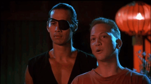 A Man With A Bandage On His Eye Is Touching Another Man 'S Face In A Dark Room GIF