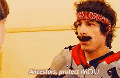 a man with a bandana on his head is talking to another man while wearing armor .